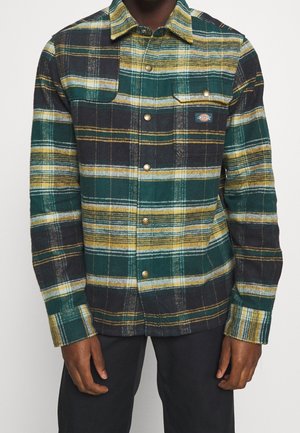 Homme portant une chemise à carreaux vert, jaune et noir avec poche poitrine et pantalon sombre, debout devant un fond uni.