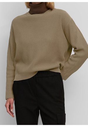 Persona che indossa un maglione beige a coste sopra un dolcevita marrone e pantaloni neri, posando con una mano sul fianco.