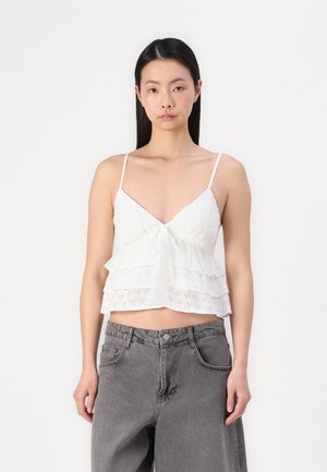 Haut sans manches blanc avec un col en V, volants superposés et détails de broderie anglaise. Associé à un jean large taille haute gris.