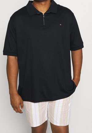 Man draagt een zwart polohemd met korte mouwen en een ritssluiting, met lichtgekleurde gestreepte korte broek, staand met één hand in zijn zak.