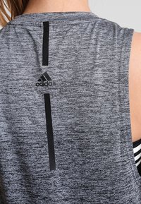 Harmaa urheilutoppi, jossa on musta pystysuora raita selässä ja musta Adidas-logo. Kankaan rakenne on teksturoitu.
