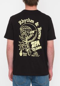 Zwarte katoenen t-shirt met een grote gele graphic op de achterkant met tekst en een ontwerp van een saxofoon, spinnenweb en muziekelementen.