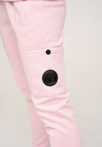 Un pantalon rose en tissu lisse présente une poche latérale avec un bouton-pression noir et un accent noir rond sur la cuisse supérieure.