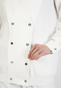 Witte geribbelde jas met metalen drukknopen, voorzien van een relaxte pasvorm en elastische zoom. De gladde textuur benadrukt het design.