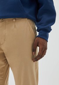 Beige, leichte Hosen mit einer glatten Textur, ausgestattet mit Seitentaschen und einem Knopfverschluss, kombiniert mit einem dunkelblauen Pullover.