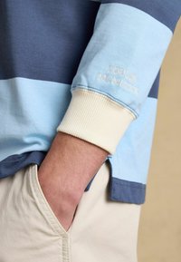 Braccio con camicia a righe blu a maniche lunghe con ricamo "Joules MCMLXXX", mano appoggiata nella tasca dei pantaloni beige su sfondo neutro.