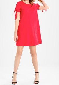 Robe rouge à manches courtes avec une coupe lâche, présentant des détails de nouage sur les manches et une texture lisse. Portée avec des escarpins noirs à bout ouvert.