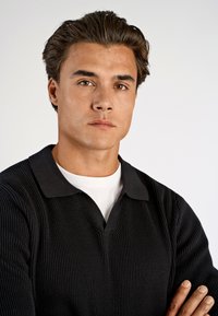 Schwarzes geripptes Poloshirt mit kontrastierendem Kragen, getragen über einem weißen T-Shirt, mit einer taillierten Passform und strukturiertem Stoff.