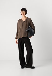 Nainen seisoo päällään ruskea pitkähiha, mustat leveälahkeiset housut, mustat kengät ja hän pitää suuren mustan clutch-laukun kainalossaan.