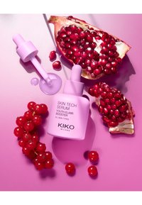 Botella de suero de vidrio rosa con cuentagotas etiquetada "KIKO SKIN TECH SERUM." Rodeada de semillas de granada y grosellas rojas sobre un fondo rosa.