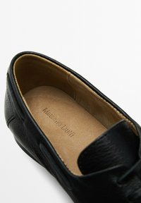 Chaussure en cuir noir avec un extérieur texturé, un intérieur lisse couleur tan et une étiquette de marque embossée. Bout arrondi et détails de design minimalistes.