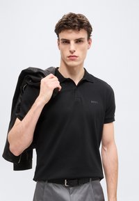 Νεαρός άνδρας με μαύρο πόλο πουκάμισο κρατώντας μαύρο δερμάτινο μπουφάν στον ώμο, σε απλό γκρι φόντο.