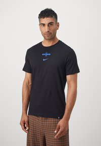 Black cotton t-shirt featuring a blue Nike logo and horizontal stripes. Short sleeves and crew neckline, paired with brown checkered shorts.