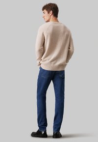 Maglione lavorato a maglia beige con orlo e polsini a costine, abbinato a jeans in denim blu. Il modello è in piedi ruotato leggermente di lato.