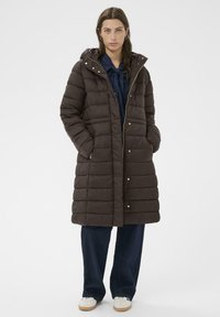 Braune Daunenjacke mit Kapuze, ausgestattet mit einem gesteppten Design, goldenen Hardware-Details und seitlichen Taschen. Wurde über einem blauen Oberteil und dunklen Hosen getragen.