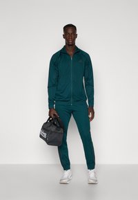 Turkizni trenažer iz gladke tkanine z črnimi detajli, jakna z zadrgo, hlače z elastičnim pasom in sivo torbo za športno opremo z belim logotipom.