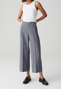 Femme portant un haut blanc sans manches, un pantalon gris large raccourci et des mocassins noirs, debout les mains dans le dos.