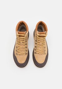 Bottes de randonnée marron clair et foncé avec une tige en daim, une semelle en caoutchouc et des lacets jaunes équipés d'œillets métalliques. Surfaces lisses et texturées.