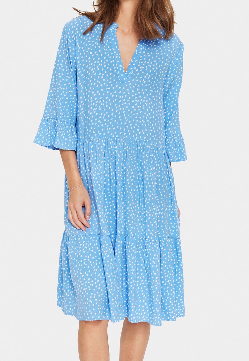 Blå prickig klänning med V-ringning, trekvartsmånga utsvängda ärmar och ett lager på lager-skirt. Tillverkad av lättviktigt tyg med en mjuk textur.