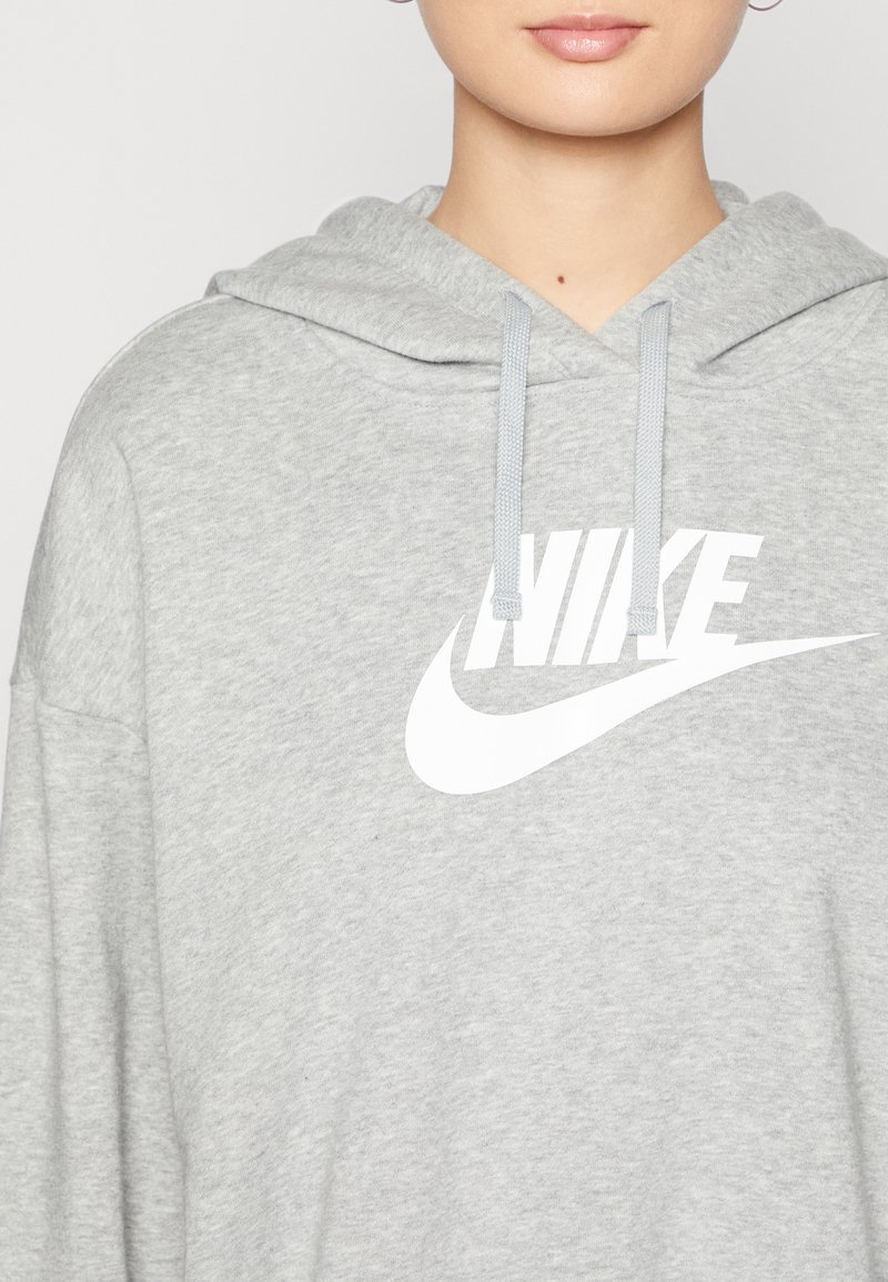 Šedá mikina s kapucí, na přední straně zdobená velkým bílým logem Nike. Materiál vypadá jemně a má mírnou strukturu.