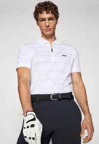 Hvit polo skjorte med glidelås krage og lys grå geometrisk mønster, paret med mørke bukser og et svart vevet belte. Hvit golfhanske i hånden.
