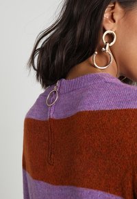 Femme aux cheveux courts et foncés porte de grandes boucles d'oreilles argentées en forme de créoles et un pull à blocs de couleurs violet et rouille avec un anneau argenté sur la fermeture éclair au dos.