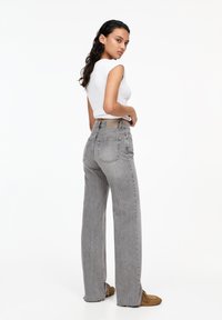 Jeans grii cu picioare largi și talie înaltă, având buzunare pe spate și un eticheta bej. Purtați cu un top alb fitted fără mâneci.