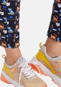 Beżowe siatkowe buty sportowe z iryzującymi akcentami i żółtymi detalami. Białe sznurowadła i teksturowana różowa podeszwa. Spodnie mają wzór z kotami.