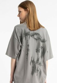 Kobieta z prostymi jasnobrązowymi włosami nosi oversize'owy szary T-shirt z czarnym abstrakcyjnym i kropkowanym wzorem na plecach na białym tle.