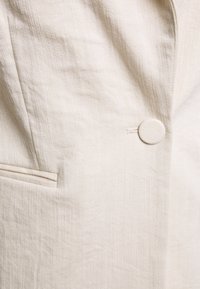 Gros plan d'un blazer en tissu beige clair avec un seul bouton recouvert et une petite poche horizontale sur le côté gauche.
