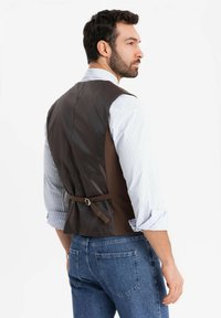 Mann in einer dunkelbraunen Weste über einem hellblauen gestreiften Hemd mit hochgekrempelten Ärmeln und blauen Jeans, der von hinten steht und zur Seite schaut.