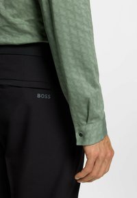 Camicia verde a fantasia con una trama geometrica, abbinata a pantaloni neri con il logo "BOSS". Maniche lunghe con polsini a bottoni.