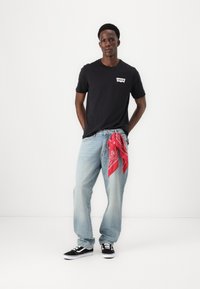Melns T-krekls ar Levi's logotipu, gaiši zili brīvi pieguloši džinsi, sarkana bandana piesieta jostasvietā un melni sporta apavi ar baltām zolēm.