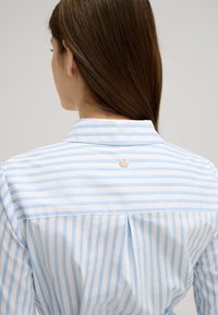 Camicia a righe blu chiaro e bianca con piega sul retro e un piccolo emblema metallico sul colletto. Tessuto liscio con un design classico.