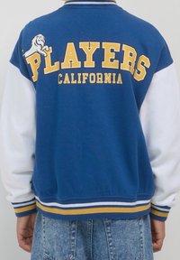 Niebiesko-biała kurtka varsity z żółtym napisem "PLAYERS CALIFORNIA" na plecach; posiada paskowane ściągacze na mankietach i dole.