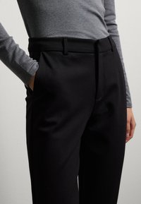 Zwarte op maat gemaakte broek met zijzakken, gemaakt van gladde stof. Heeft een recht gesneden ontwerp en een middelhoge tailleband. Grijze longsleeve bovenkant zichtbaar.
