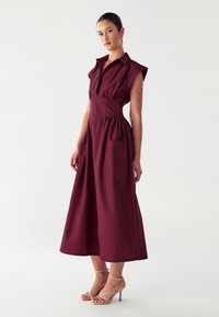 Abito midi bordeaux con maniche corte, colletto, vita arricciata e due tasche anteriori. Tessuto liscio con una forma strutturata.