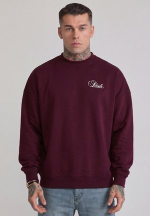 Uomo con tatuaggi al collo e alla mano che indossa una felpa bordeaux con il logo bianco "Siksilk" e jeans azzurri chiari su uno sfondo uniforme.