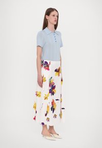 Lichtblauwe polo met korte mouwen en knopen, gecombineerd met een witte maxi-rok met bloemenprint en kleurrijke bloemen in een vloeiende ontwerp.