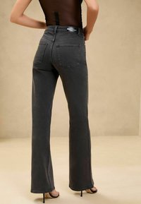 Personne portant un jean droit taille haute gris foncé et un haut noir transparent, debout les mains sur les hanches devant un fond beige.