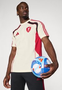 Cremefarvet og rød t-shirt med korte ærmer og LFC-logo, med sort kant og tre stribede detaljer. Modellen holder en blå og hvid fodbold.
