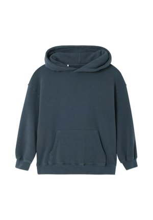 Kind staat, draagt een donkere hoodie, lichtgrijze joggingbroek en blauwe sandalen, met haar in een knot tegen een effen witte achtergrond.