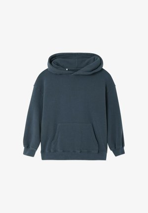 Kind staat, draagt een donkere hoodie, lichtgrijze joggingbroek en blauwe sandalen, met haar in een knot tegen een effen witte achtergrond.