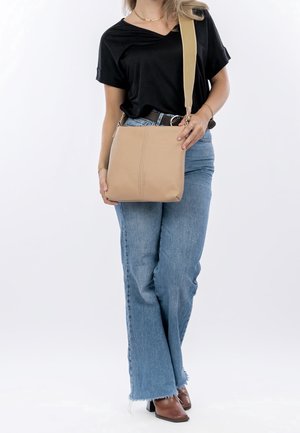 Borsa a tracolla in pelle color tan con una finitura liscia e un design cucito. La tracolla è ampia e beige, in abbinamento a jeans blu scuro e una maglietta nera.