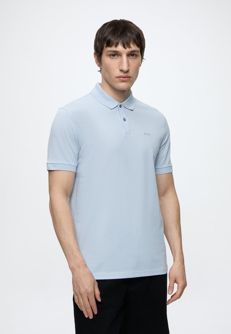 Ung mand iført lyseblå kortærmet polo-shirt med knapper, stående mod en ensfarvet hvid baggrund, vendt mod fronten med neutralt udtryk.