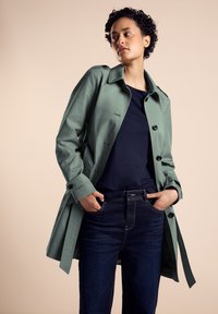 Ljusgrön trenchcoat med avslappnad passform, knappstängning och bälte. Bärs över en marinblå topp och mörka jeans. Slät tygtextur.