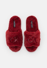 Chaussons rouges en peluche avec un tissu doux, design ouvert à l'avant et logo "CK" brodé sur la bande. Texture douce pour un confort amélioré.