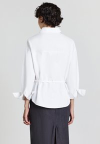 Person mit kurzen lockigen Haaren, die eine weiße Bluse mit langen Ärmeln und einem Taillengürtel sowie einen dunkelgrauen Rock trägt, von hinten gezeigt.