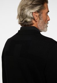 Profil av en mogen man med grått hår och skägg, iklädd en svart tröja med ett subtilt texturerat mönster på ryggen.