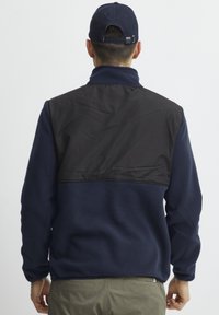 Giacca in pile blu navy con un pannello superiore in nylon nero. Presenta un colletto alto, maniche lunghe e una texture liscia. Design semplice, senza motivi.